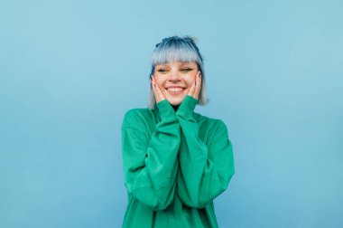 Mavi saçlı neşeli kız yeşil bir arka planda duruyor, yüzünde bir gülümsemeyle ve mutlu bakışlarla..