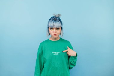 Tatminsiz, renkli saçlı hippi kız mavi arka planda duruyor ve kendini işaret ediyor ve kameraya bakıyor ve ıskalıyor.