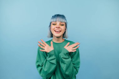 Renkli saçlı neşeli kız mavi bir arka planda duruyor ve mutlu bir yüz ile kameraya bakıyor.