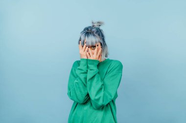 Scared hipster girl in a green sweatshirt and with blue hair stands on a blue background and covers her face in fear, looking at the camera.