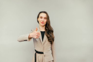 Takım elbiseli pozitif kadın ofis çalışanı bej arka planda duruyor, kameraya bakıyor ve gülümsüyor ve baş parmağını kaldırıyor.