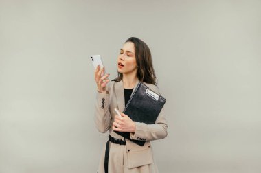 Takım elbiseli bir kadın işçi bej bir arka planda elinde tabletle duruyor ve ciddi bir yüzle akıllı telefon kullanıyor..