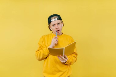 Pensive guy student in headphones with a notebook isolated on a yellow background, looking at the camera with a serious face and biting a pen.