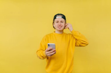 Şapkalı ve günlük kıyafetli genç adam sarı bir arka planda elinde akıllı bir telefonla duruyor ve yüzünde bir gülümsemeyle kameraya bakıyor, kulaklıkla müzik dinliyor.