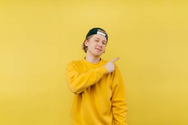 Şapkalı ve kazaklı mutlu adam sarı arka planda duruyor ve yüzünde bir gülümsemeyle parmağını yana gösteriyor..