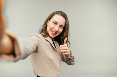 Takım elbiseli mutlu iş kadını bej arkaplanda özçekim yapıyor. Yüzünde gülümseme ve hareketler sergiliyor..