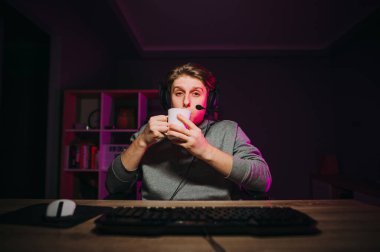 A handsome young man in a headset sits at night on a computer at home with a cup of tea in his hands and looks at the camera with a serious face.