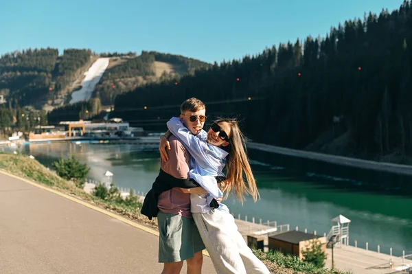 在一个阳光明媚的夏日 在高山和湖泊的背景下 一对快乐的情侣正在一个山庄里散步 玩得很开心 看着相机 微笑着 — 图库照片