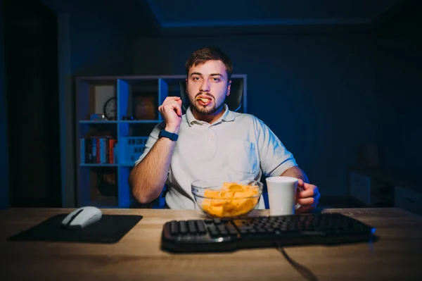 An adult man in a white T-shirt works at night at the computer and eats chips and tea sitting in a dark room with blue light.