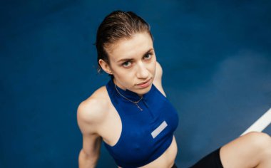 Yağmurda antrenman yaparken ıslak bir spor sahasında uzanmış mavi arka plandaki kameraya bakan spor elbiseli güzel bir kadının portresi.