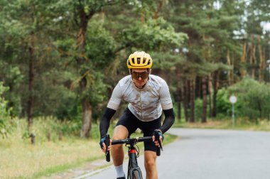 Spor giyimli bir erkek bisikletçi ciddi bir yüzle asfalt bir yolda şehir dışına çıkıyor..