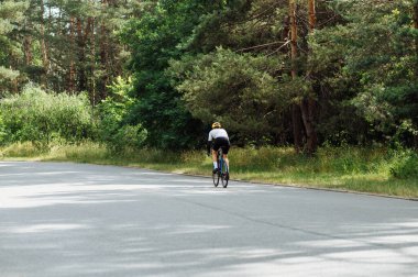 Spor giyimli erkek bisikletçi, ormanın içinde asfalt bir yolda gidiyor..