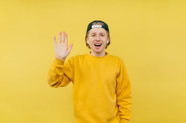 Renkli kazaklı ve şapkalı neşeli genç bir adam avucunu kameraya uzatıp yüzünde bir gülümsemeyle kameraya bakıyor.