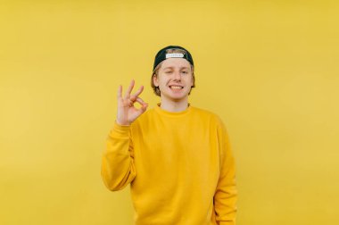 Şapkalı neşeli genç adam ve parlak yumurta geyik kıyafetleri sarı arka planda duruyor, yüzünde bir gülümsemeyle kameraya bakıyor ve bir jest gösteriyor.