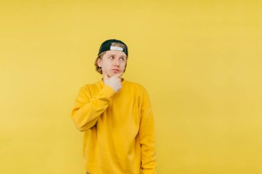 Üzerinde eşofman altı ve şapka olan düşünceli bir adamın portresi sarı bir arka planda duruyor ve başka tarafa bakıyor ve düşünüyor..