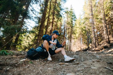 Dağlarda erkek yürüyüşçü elinde bir şişe suyla patikanın kenarında oturmuş, dinleniyor ve akıllı telefon kullanıyor.