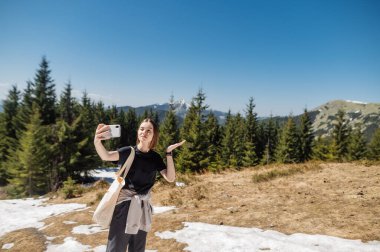 Güzel manzaralı bir arka planda, akıllı bir telefondan selfie çeken, günlük giysiler içindeki çekici bir kadın.