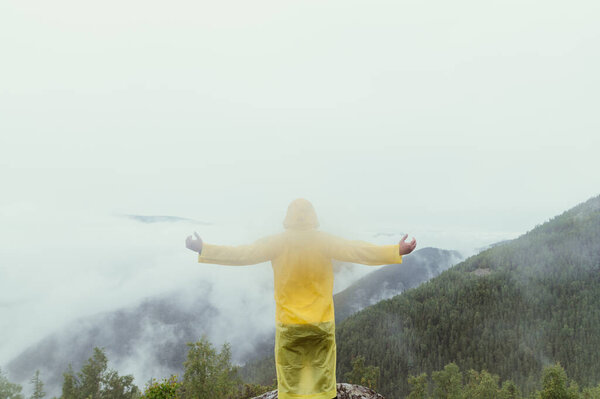 A man in a yellow raincoat stands on top of a mountain on a rock and looks at a beautiful misty view with his hands raised.