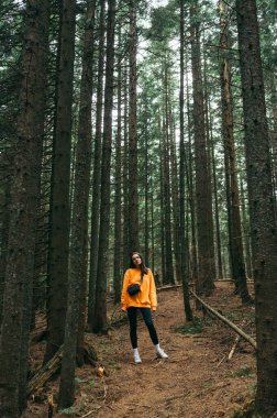 attractive woman in casual clothes stands on a path in a mountain forest and poses for the camera with a smile on her face.