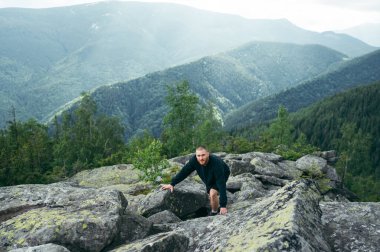 Active young man in casual clothes is climbing a rock in the mountains at the top and looking at the camera.