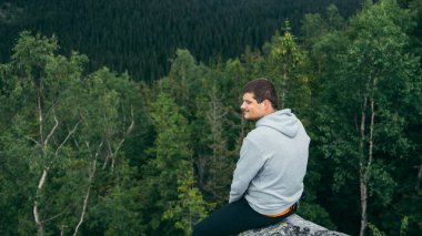 Positive young male tourist sits on a rock in the mountains and looks away with a smile on his face.
