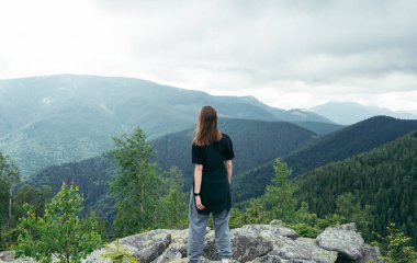 Photo of a female tourist from the back standing on a rock on top of a mountain and looking at a beautiful landscape. Active recreation hiking in the mountains.