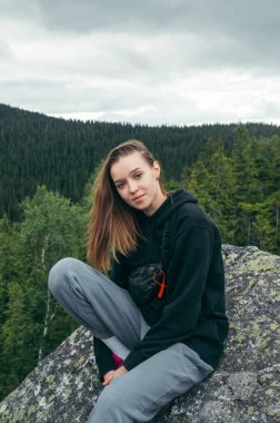 Portrait of an attractive female tourist in casual clothes sitting on a rock in the mountains and posing for the camera against the background of the Carpathians. Vertical