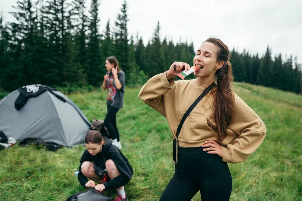 Woman hiker eating a protein bar in the mountains against the background of friends hikers and campers during a hike.