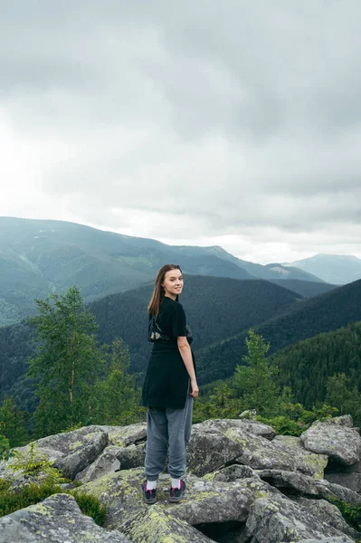Positive woman in casual clothes stands on a rock in the mountains during a hike and poses for the camera against the background of Carpathian views with a smile on her face. Vertical