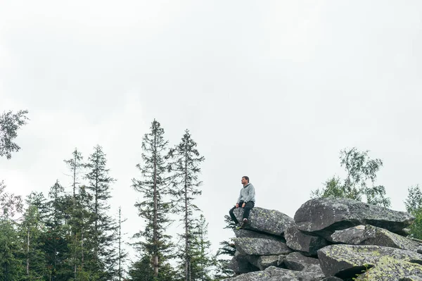 A male tourist in casual clothes sits on a rock on top of a mountain and looks at the views. Active recreation hiking in the mountains.