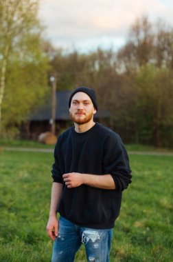Handsome stylish young man in casual clothes and a hat and with a beard is standing in the park and posing for the camera.