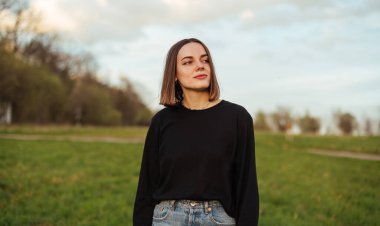 Stylish beautiful lady in dark clothes standing outdoors in field at sunset, looking away with serious face and posing for camera.