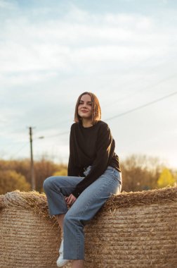 Attractive girl in casual clothes poses for the camera on a hay bale in a field with a smile on her face. Vertical