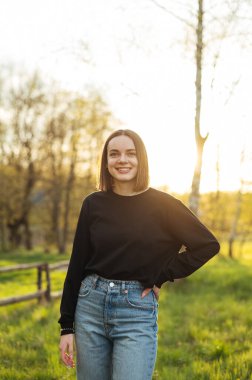 Portrait of an attractive woman in stylish casual clothes on the street posing for the camera with a smile on her face against a sunset background with trees.