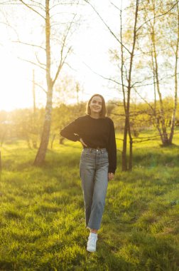 Full length portrait of an attractive woman in casual clothes standing outdoors in the park and looking at the camera with a smile on her face.