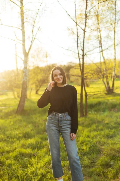 Positive woman in casual clothes posing for the camera in the park at sunset, looking at the camera with a smile on her face.