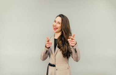 Dişinde diş teli olan ve bej takım elbiseli neşeli bir kadın bej bir arka planda duruyor ve yüzünde bir gülümsemeyle parmaklarını kameraya gösteriyor.