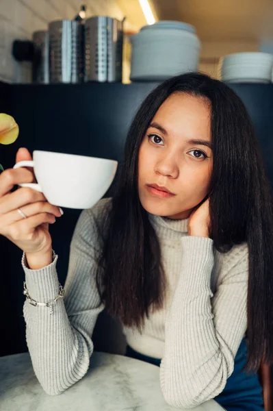 Portrait of a young Hispanic woman with a cup of coffee in her hands sitting in a cafe at the table and looking at the camera with a serious face.