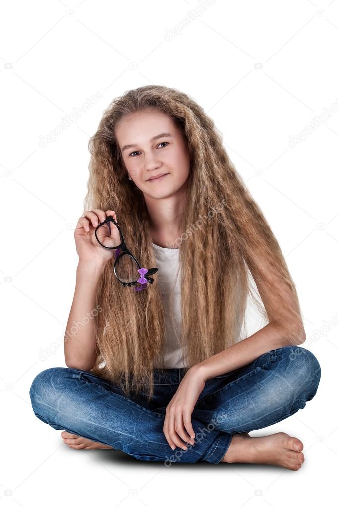 Cute smiling little teenage girl with long hair sitting on the floor with crossed legs, isolated on white background