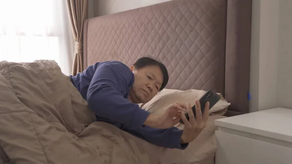 Awake elderly old Asian woman, people waking up, using a smartphone on social media internet and sleeping on bed in bedroom at home. Lifestyle in technology device concept in morning.