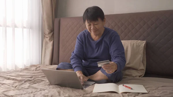 幸せな高齢者のアジアの女性は 人々のオンラインショッピング 自宅の寝室でベッドの上のコンピュータノートパソコン ビジネス文書でお金を支払うためにクレジットカードを使用して ライフスタイル — ストック写真