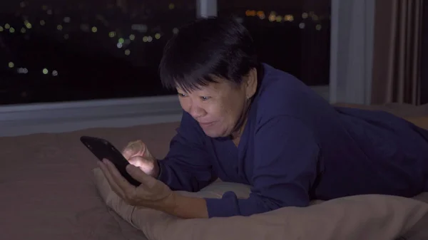 Happy elderly old Asian woman, people using a smartphone on social media internet and sleeping on bed in bedroom at home. Lifestyle on late night in technology device concept. Insomnia.