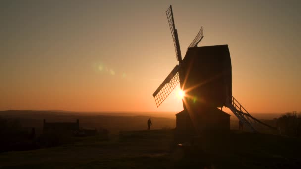 日陰で犬を歩く無名の人々と日没時に伝統的な木製の風車の後ろの地平線に太陽が沈む — ストック動画