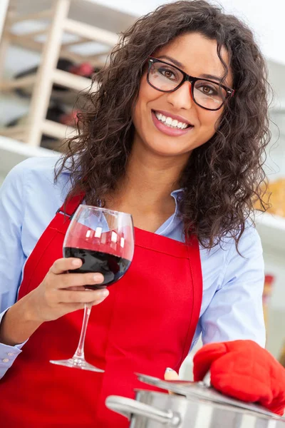 美しい幸せなヒスパニックラティーナ若い女性または女の子とともに完璧な歯を身に着けています赤いエプロン飲むAガラスの赤ワインと料理で彼女の台所で彼女の家 — ストック写真