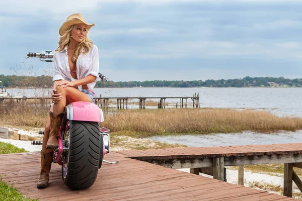 セクシーブロンド女の子若いです女性身に着けていますピンクビキニ ジーンズショートパンツ カウボーイブーツと帽子座っています上のピンクチョッパーオートバイによってA湖 — ストック写真