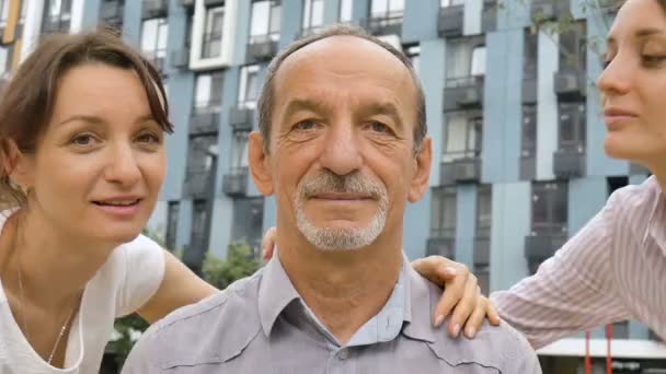 Family portrait of senior father and two adult daughters kissing him outdoors on modern building of new residential complex, fathers day celebration concept — Stock Video