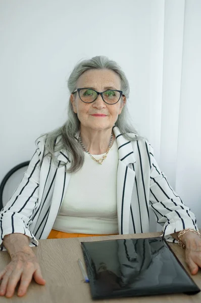 Старшая седовласая деловая женщина в полосатой куртке в очках работает в своем офисе, сидя за столом. Счастливого выхода на пенсию, работы и труда — стоковое фото