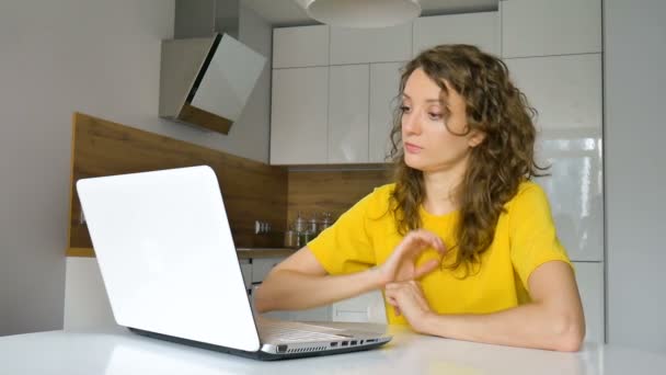 Mujer joven con el pelo rizado y camisa amarilla está trabajando desde casa usando su computadora portátil en la mesa de la cocina en su apartamento, trabajo remoto, freelance — Vídeos de Stock