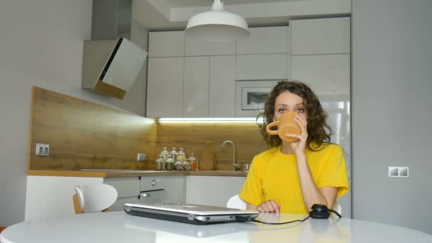 Tired young woman with curly hair and yellow shirt is working from home using her laptop at the kitchen table in her apartment, remote work, freelance, burnout syndrome — Stock Video