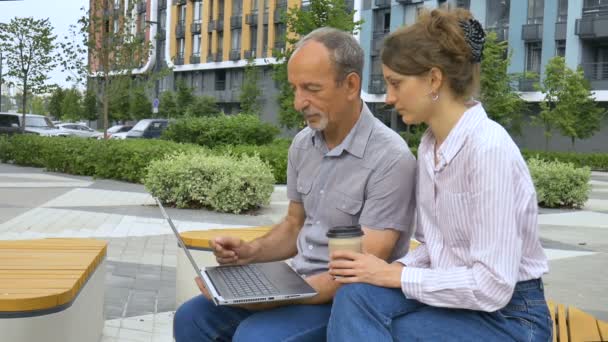 Jovem funcionário está ensinando seu colega mais velho como usar laptop e software corporativo sentado no banco fora do escritório, coordenar o estágio, parceria de educação. — Vídeo de Stock